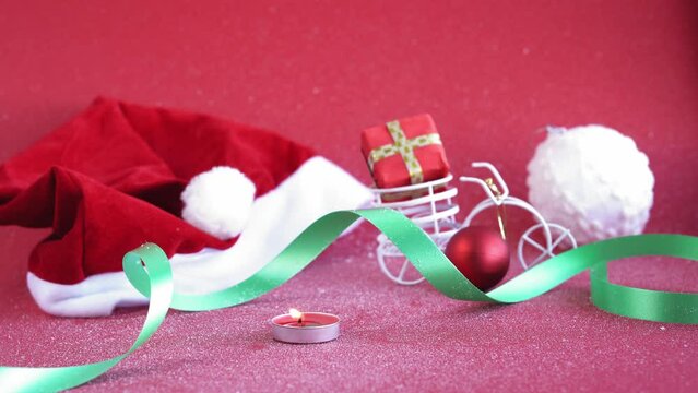 Xmas, winter, new year concept - red Christmas background covered with white snow Santa hat with burning candle and toy bicycle with figuratively gift in trunk. Flatly, top view, overhead looped video
