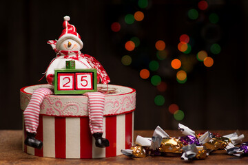 A snowman figure is sitting on a striped decoupage box and holding a smaller box with two cubes forming a number twenty-five. A bunch of parlour candies are lying beside.