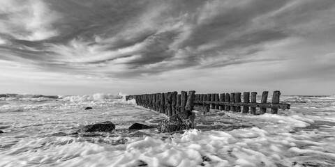Black and white picture of wavebreakers disappearing in the sea and some rocks in the foreground under a clouded sky , at the coast of Zeeland, The Netherlands