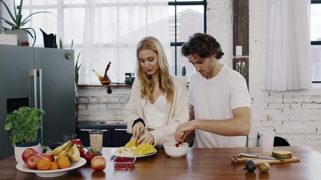 Young attractive couple cuts fruits together and prepare a healthy fruit-bowl, porridge-bowl, acai-bowl in modern kitchen while having fun