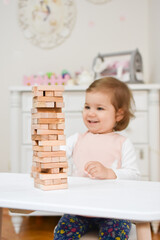 Cute adorable baby girl playing table game in her room at home. Beautiful kid and jenga blocks. Child playing brain games. White scandinavian interior for children's room. Nursery with nordic stylish.