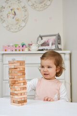 Cute adorable baby girl playing table game in her room at home. Beautiful kid and jenga blocks. Child playing brain games. White scandinavian interior for children's room. Nursery with nordic stylish.