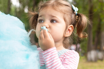 Cute adorable baby girl eating blue cotton candy outdoor. Funny child joining cotton candy in park. Pretty toddler girl holding desert in garden.
