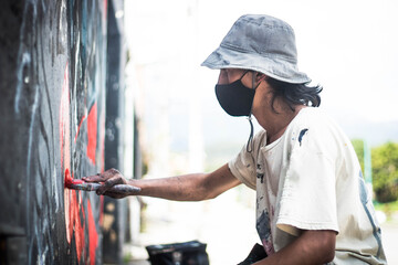 A handsome graffiti artist in a face mask bucket hat and paint-stained clothes painting with a paintbrush on a wall