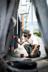 A handsome graffiti artist in a face mask bucket hat and paint-stained clothes painting with a paintbrush on a street wall