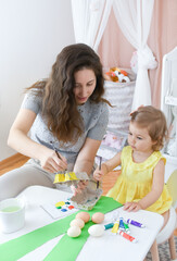Cute adorable baby girl paint colorful eggs with mother. Happy family prepare for Easter. Happy easter! Child and mom in nursery. Kid doing craft in children's room with a white Scandinavian interior.