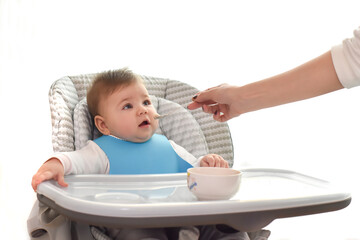Young mother feeding her cute adorable baby, whom sitting on the grey highchair, with a spoon. Family, first food, child, eating, parenthood and motherhood concept