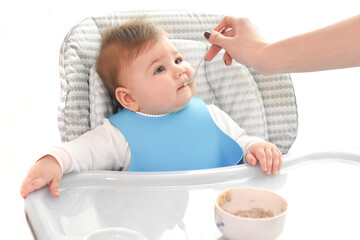 Young mother feeding her cute adorable baby, whom sitting on the grey highchair, with a spoon. Family, first food, child, eating, parenthood and motherhood concept