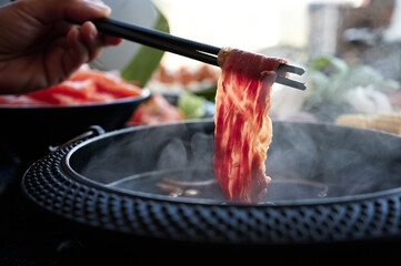a hand holds marbled wagyu beef with chopstick above a steamy hot pot in a buffet japanese restaurant