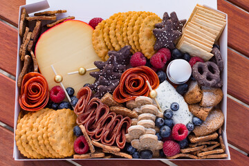 Top view of a boxed grazing board of charcuterie, crackers, berries, cookies, and cheeses on a wood table