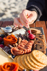 Woman's hand selecting a red raspberry from a grazing board filled with charcuterie, tapas, crudites, nuts, crackers, cheeses, and food sitting on a wood table with blurry sand in background 