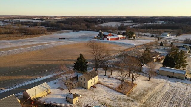 Establishing shot of Midwestern countryside in winter.  Aerial view of  rustic road, farm houses, farms along the street. Agricultural field  covered in first snow, sunny, soft sunlight at sunset 