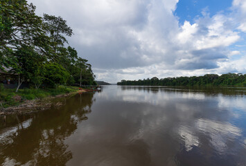 Fototapeta na wymiar Long River Through Jungle Scenery In Brokopondo Suriname. Lush Green Trees Shadowed By Overcast White Clouds In South America.