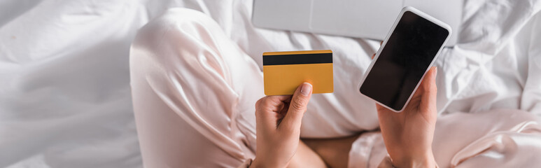 cropped of woman sitting in bed with smartphone, credit card and laptop at morning, banner