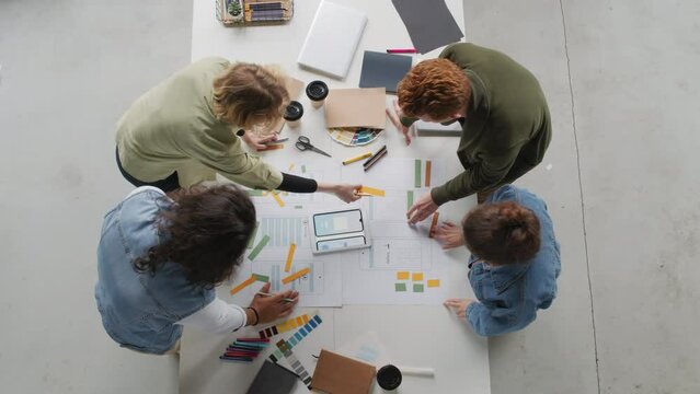 Top view of team of young app developers standing around table in meeting room and working on interface for new project