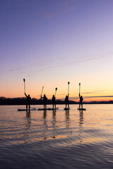 Fototapeta na wymiar Five friends on stand up paddle board (SUP) on a flat quiet winter river at sunset raising his paddles up. Vertical image