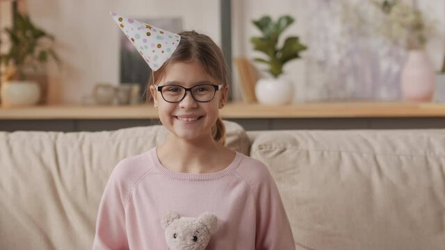 Tilting-up portrait of pretty little girl in glasses and colorful festive cap sitting on sofa in living room hugging white stuffed hare from present box smiling at camera