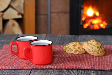 A two red mugs of black tea and cookies are on a wooden table against fireplace with burning firewood in a rural house. 