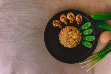 Shrimp fried rice in a black plate with spring onion, egg and cucumber all on the wooden table. top view.
