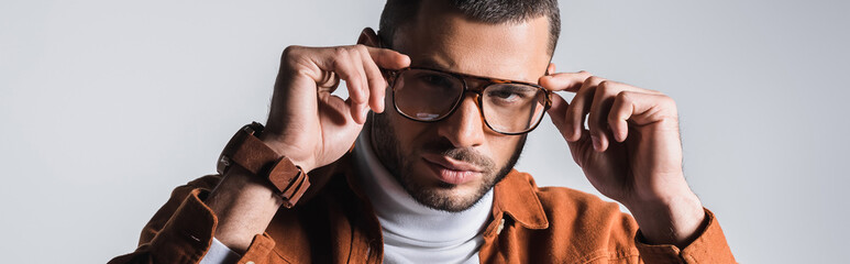 Stylish man adjusting eyeglasses and looking at camera isolated on grey, banner - obrazy, fototapety, plakaty