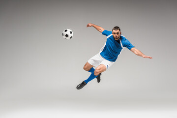 Fototapeta na wymiar Sportsman looking at camera while jumping near football on grey background