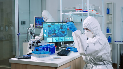 Fototapeta na wymiar Chemist in coverall checking blood samples taking notes on computer in equipped laboratory. Team of doctors examining vaccine evolution using high tech researching diagnosis against covid19 virus