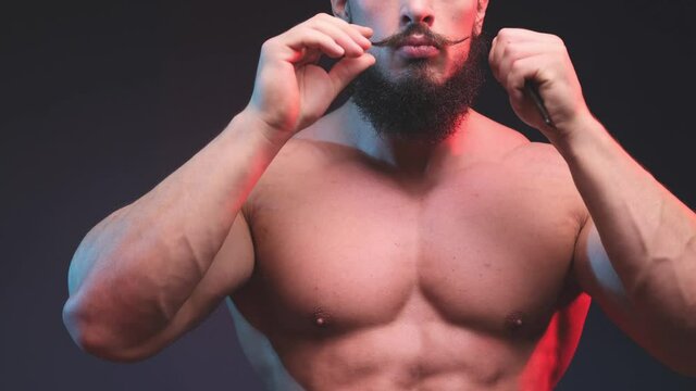 Handsome and muscular hipster guy with naked body poses in dark background combing his beard and looking at camera. Funny caucasian athlete with stylish moustache and haircut.