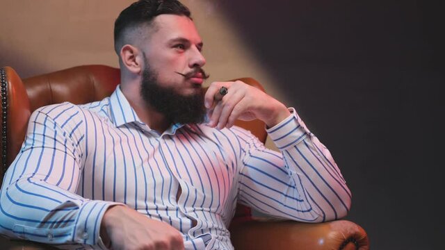 Stylish and handsome macho with muscular build and modern haircut sits on leather and luxurious armchair smoking cigar in dark room. Succesful elegance bodybuilder with cigar and fashionable beard