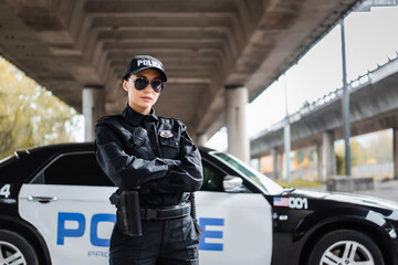 confident policewoman with crossed arms looking at camera near patrol car on blurred background on...