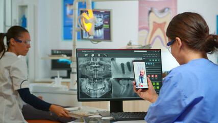 Nurse having video call with expert stomatologic medic while doctor is working with patient in background. Stomatologist assistant listening dentist using mobile webcam sitting on stomatological chair - obrazy, fototapety, plakaty