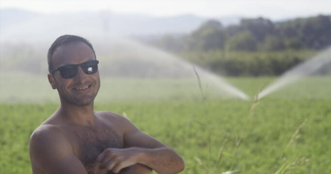 Agriculture worker with no shirt on a green field with irrigation system. Water sprinklers in the background
