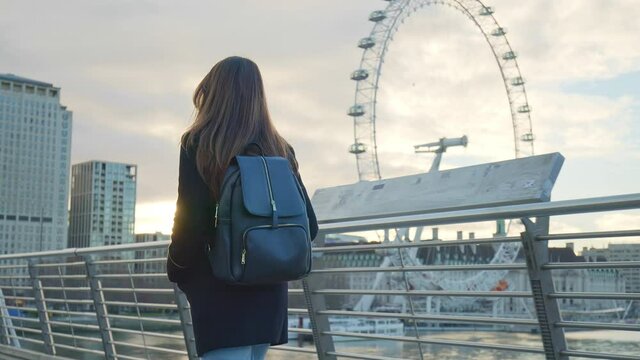 A girl tourist with backpack walking on Millennium Bridge, London in lockdown. A female in a coat during winter with no people walking in front of most famous London landmarks in slow-motion in 4K.