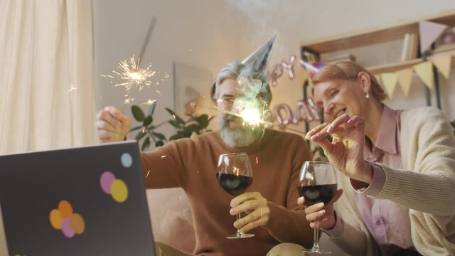 Medium slow-motion waist-up footage of cheerful middle-aged couple celebrating birthday at home with red wine and sparklers having video chat with family via laptop during quarantine