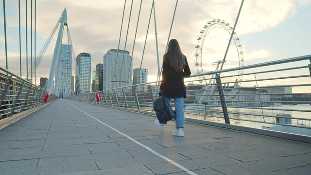 A girl tourist with backpack walking on Millennium Bridge, London in lockdown. A female in a coat during winter with no people walking in front of most famous London landmarks in slow-motion in 4K.
