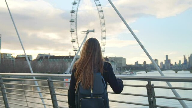 A girl tourist with backpack walking on Millennium Bridge, London in lockdown. A female in a coat during winter with no people walking in front of most famous London landmarks in slow-motion in 4K.