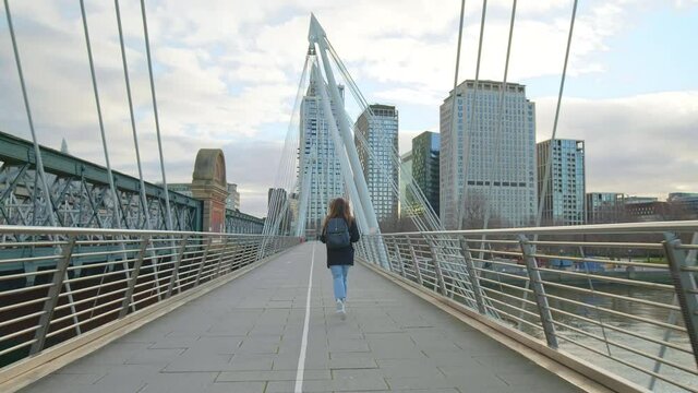 A girl tourist with backpack running on Millennium Bridge, London in lockdown. A female in a coat during winter with no people running in front of most famous London landmarks in slow-motion in 4K.