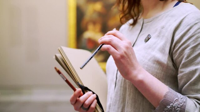 Unrecognizable female artist draws simple sketches in paper notebook with brown pages while visit art museum and enjoying ancient masterpiece of painting 