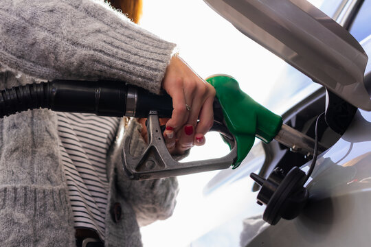 A young woman with painted nails and a nice sweater, fills up her car with gasoline by helping herself with two hands. Image taken from a low angle