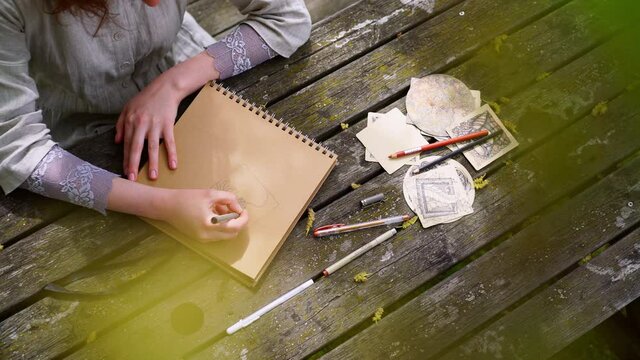 Close-up art supplies on wooden worktop and above view faceless woman draw in sketchbook with pencil while sitting at table in sunny spring garden of historical city park