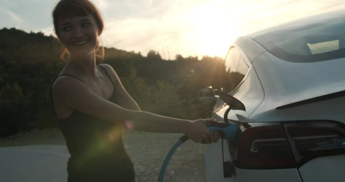 Girl having trouble removing a charging cable from her electric car. Handheld video during sunset