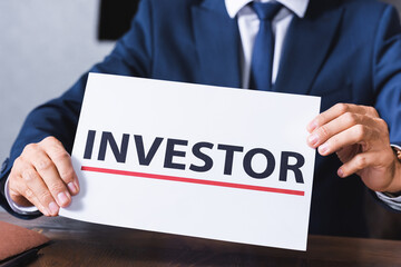 Cropped view of businessman holding sign with investor lettering in meeting room on blurred background.