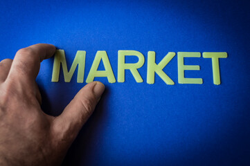 Human fingers holding the word Market written with plastic letters on blue paper background, concept