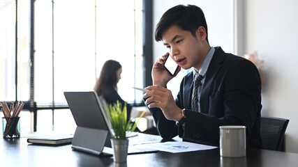 Businessman in suit talking on mobile phone and working with tablet computer while sitting in office room.