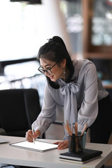 Portrait of cheerful woman in eye glasses standing at her workspace and using digital tablet.
