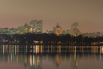 Fototapeta premium Scenic night cityscape of Obolon neighborhood with modern skyscrapers. Buildings reflected in the tranquil water of Dnieper River. Embankment with renovated Natalka Park. Kyiv, Ukraine
