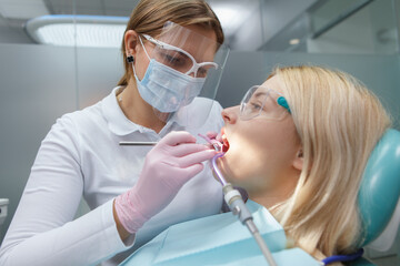 Female dentist wearing protective face mask, treating teeth of female patient
