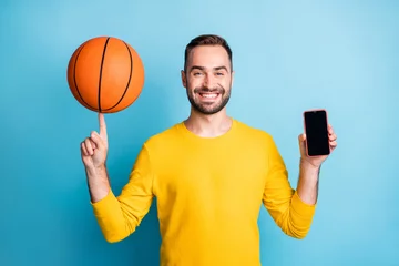Foto op Plexiglas Photo of young handsome positive good mood smiling man hold phone and basketball ball on finger isolated on blue color background © deagreez