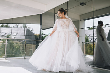 A smiling bride with curly hair in a white dress with a long veil dances and whirls. Wedding portrait in the morning at the hotel.