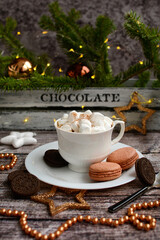 Vertical New Year composition. Coffee with mashmelou in a white cup, macarons, brown beads, stars on the background of glowing garlands. Closeup