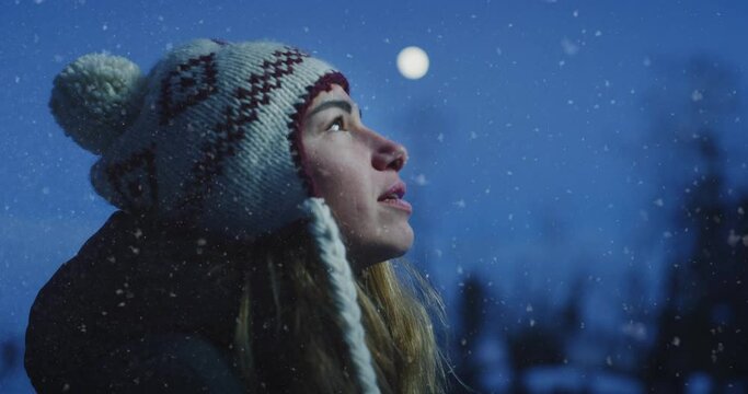 Cinematic shot of happy smiling young woman in warm hat is breathing fresh air and enjoying winter evening with full moon light while snow falling. Concept of winter holidays,freedom,happiness, nature